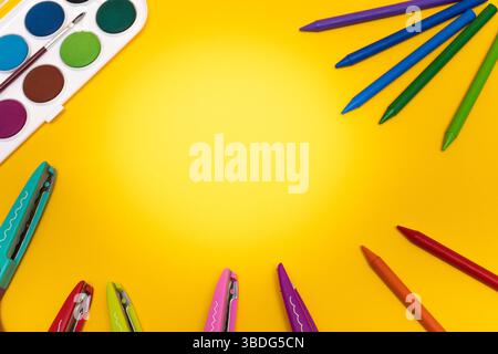 Flat lay of colorful art tools including crayons, scissors, and watercolor set on a vivid yellow background, arranged in a creative border with blank Stock Photo