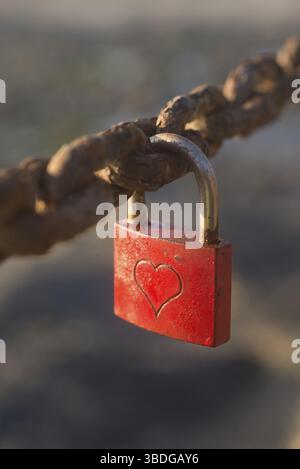 A small lock, painted red, with a heart engraved on it, is locked on to a rusty chain Stock Photo
