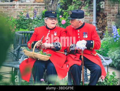 Two Chelsea Pensioners in their red uniforms sit on a bench and chat at Chelsea Flower Show, London, UK Stock Photo