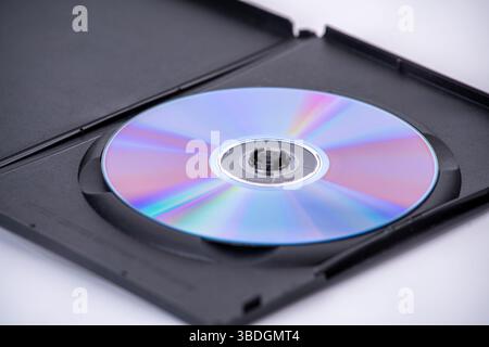 An optical disc with reflective surface is placed inside black plastic case, partially open, on white background. disc displays spectrum of colors Stock Photo