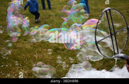 Colorful soap bubbles floating in the air with children playing in the background. Outdoor fun on a grassy field, creating giant bubbles with a wand. Stock Photo