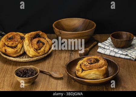 Homemade delicious raisin cinnamon cake roll in wooden plate and in bamboo tray with dried raisin in wooden cup, spoon, honey dipper, bowl and table c Stock Photo