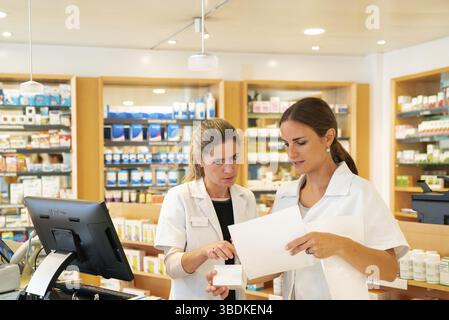 Female pharmacist consults with her assistant about a medical prescription with a young trainee pharma assistant in the background Stock Photo