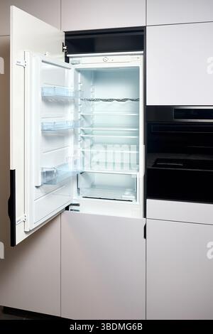 Vertical shot of kitchen interior with beige wooden furniture and modern appliances. Opened built integrated refrigerator with opened door empty shelv Stock Photo