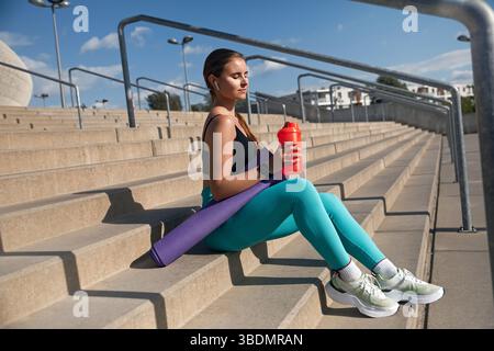 A young woman sits on the steps outdoors, holding a red water bottle while resting after her workout. Stock Photo