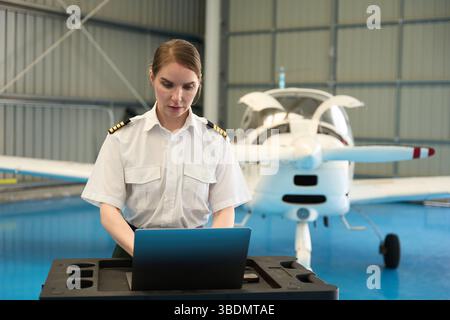 Female pilot working on laptop in aircraft hangar with small plane Stock Photo