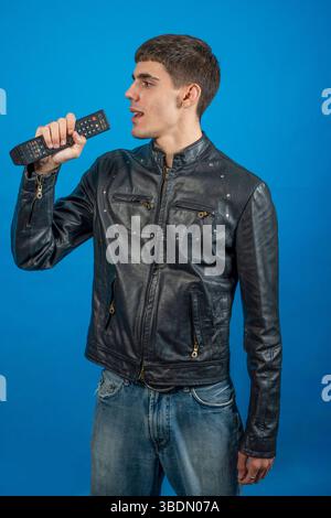 Young man using remote control as microphone, singing and having fun on blue background Stock Photo