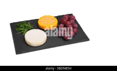 Two cheesy round heads of brie with grapes and arugula on a black slate board. A snack. Isolated cheese board on a white background. Stock Photo
