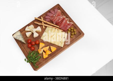 Cheese wooden board on white table. Different types of cheese, chili, jamon, sausage, olive, tomato, microgreens, knife, breadsticks. Stock Photo