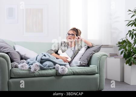 Woman in pain holding a hot water bottle on her stomach while lying on a couch at home, wearing smart glasses and interacting with virtual screen to f Stock Photo