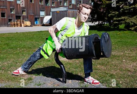 A lifelike sculpture depicting a maintenance worker in a fluorescent safety vest tending to a public trash bin on a grassy area, with apartment buildi Stock Photo