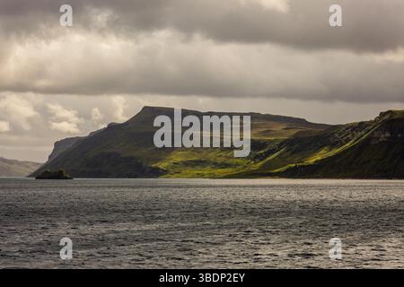Late sunlight illuminates green coastal slopes and sea cliffs on the Isle of Skye, Scotland, with a dark sky and small islet in foreground. Stock Photo
