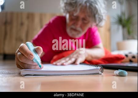 Elderly man lying on the floor and using a green marker to write or draw in a spiral notebook. Creative indoor activity, close-up of hand, concept of Stock Photo
