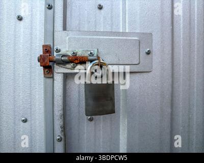 Photograph of an old padlock that is securing a rusty single eye padbolt door locking system on an old galvanised shed in a residential garden. Stock Photo