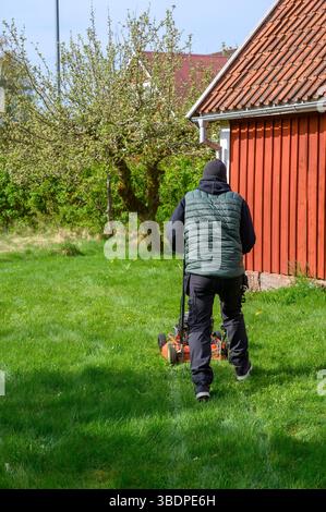 A person in dark clothing operates a lawn mower on a vibrant green lawn next to a charming red wooden house. The sun shines brightly, indicating a ple Stock Photo