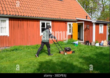 A person in dark clothing operates a lawn mower on a vibrant green lawn next to a charming red wooden house. The sun shines brightly, indicating a ple Stock Photo