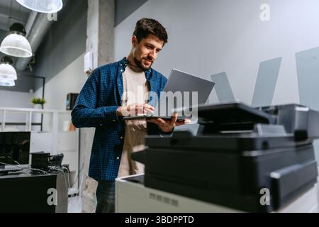 Man using laptop in modern office space while standing next to printer during working hours Stock Photo