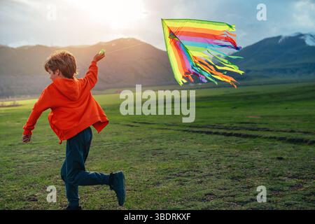 Happy boy runs with kite on green field on mountains background at sunset Stock Photo