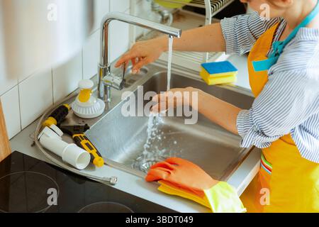 Aproned woman in gloves washes her hands at a stainless sink as plumbing tools lie nearby, finishing cleanup after fixing a kitchen drain clog. Stock Photo
