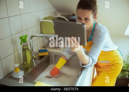 Woman in striped shirt and yellow apron uses a tablet while cleaning a soapy stainless steel sink, multitasking in a bright kitchen. Stock Photo