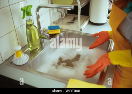 Person in orange gloves and a yellow apron stands at a soapy sink with cleaning tools, likely facing a kitchen drain clog or plumbing issue. Stock Photo