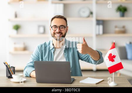 Happy young guy sitting at table with flag of Canada, using laptop computer, showing thumb up gesture at home Stock Photo