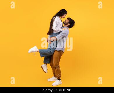 Loving indian guy lifting his girlfriend and holding her on yellow studio background, profile side view, free space banner Stock Photo