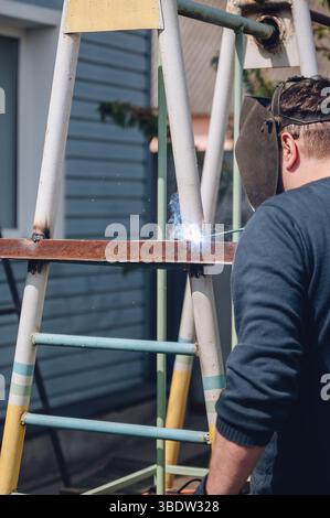 A male welder in a welding mask welds metal with a welding machine on a sports ground Stock Photo