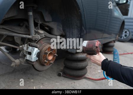 Mechanic uses an impact wrench to service a lifted car, showing exposed brake components and pneumatic air jack system. Stock Photo