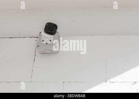 Surveillance camera mounted on a white tiled wall, slightly dusty and positioned for overhead monitoring in a bright environment. Stock Photo