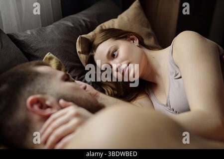 Couple lying on bed, surrounded by soft pillows, woman gazing intently at man, creating a serene and intimate moment, capturing soft light Stock Photo