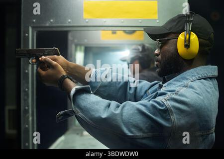 Man wearing headphones aiming pistol at indoor shooting range with focus and precision, another person adjusting equipment in background Stock Photo