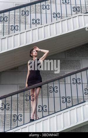 A woman in a black dress poses on a staircase with decorative railings Stock Photo