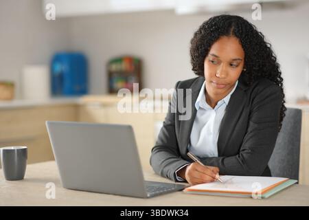 Black executive working online taking notes in a notebook and checking laptop at home Stock Photo