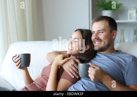 Happy couple drinking tea looking away sitting on a couch in a house interior Stock Photo
