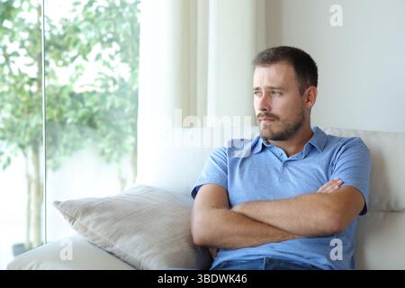 Pensive sad man looking through a window sitting on a couch at home Stock Photo