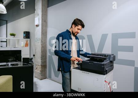 Man operates a printer in a modern office environment during daylight, focused on completing tasks efficiently and professionally Stock Photo