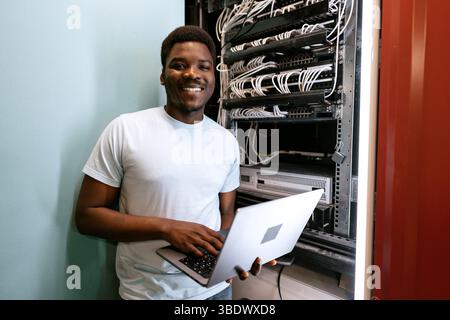Young professional managing servers with laptop while smiling in a data center during daytime Stock Photo