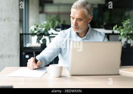 Smiling 40s male business man accountant analyst holding documents, work at laptop computer doing online trade market tech research. Focused businessm Stock Photo