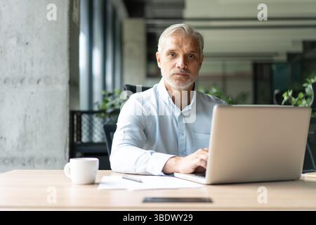 Happy mature European business man ceo trader using computer, typing, working in modern office, doing online data market analysis, thinking planning t Stock Photo
