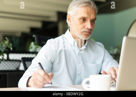 Smiling 40s male business man accountant analyst holding documents, work at laptop computer doing online trade market tech research. Focused businessm Stock Photo