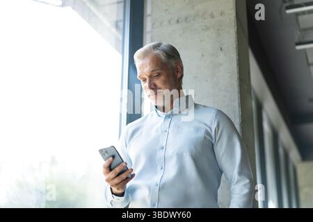 Smiling happy confident mid aged male company ceo executive holding cellphone standing in office using business mobile apps technology financial onlin Stock Photo