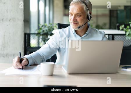 Smiling 40s male business man accountant analyst holding documents, work at laptop computer doing online trade market tech research. Focused businessm Stock Photo