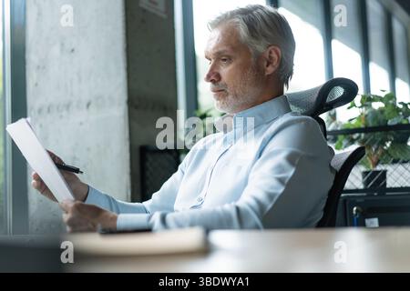Smiling 40s male business man accountant analyst holding documents, work at laptop computer doing online trade market tech research. Focused businessm Stock Photo