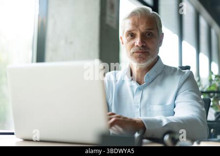 Happy mature professional business man looking at camera sitting at work desk using computer. Portrait of smiling male company worker employee, projec Stock Photo