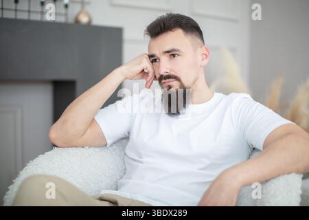 A sad and thoughtful young man with a beard. He is wearing a white shirt and leaning on his right hand. Stock Photo