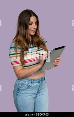 Smiling young woman using a tablet with stylus pen, working or studying on a purple background Stock Photo