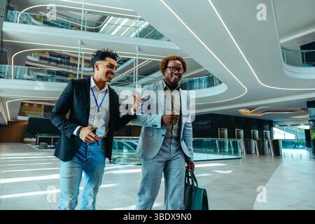 Two cheerful businessmen walk through a modern office lobby, engaging in friendly conversation. The sleek architecture of the office space highlights Stock Photo
