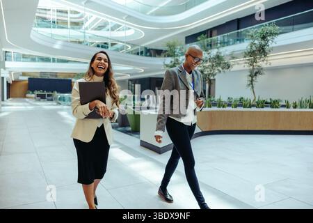 Two professionals, one Indian and one African, walk through a modern office laughing and enjoying conversation. The environment is bright and contempo Stock Photo