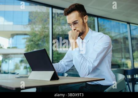 A worried businessman is deeply focused as he works on a tablet in a contemporary office environment. The large windows provide a glimpse of the outsi Stock Photo